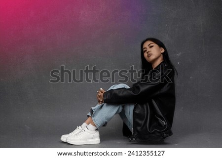 Confident young Asian girl sitting on floor in black jacket with arm around her knees, pink glow on her side. On gray background, Asian model in modern clothes poses on the floor with a pink light