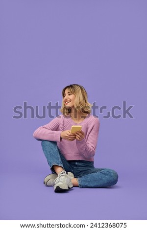 Happy relaxed gen z blonde young woman model holding smartphone looking away at copy space, smiling girl using mobile apps on cell phone sitting isolated on purple background with cellphone. Vertical