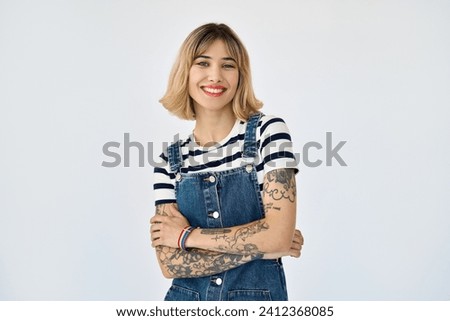 Happy pretty gen z blonde young woman, cute girl with short blond hair tattoos wearing striped t-shirt denim dress rainbow bracelet looking at camera standing isolated on white background. Portrait. Royalty-Free Stock Photo #2412368085