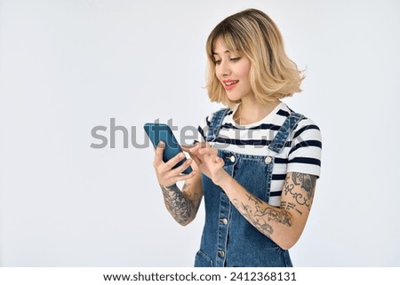 Generation z blonde young woman using smartphone isolated on white background. Cool gen z hipster girl with tattoos wearing striped t-shirt and denim dress holding mobile cell phone buying online. Royalty-Free Stock Photo #2412368131