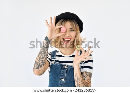 Happy pretty gen z blonde young woman, smiling hipster girl with short blond hair tattoos wearing striped t-shirt and denim dress having fun holding macaroons standing isolated on white background.
