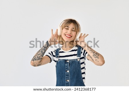 Happy funky gen z blonde young woman, cool girl with tattoos wearing striped t-shirt denim dress and headphones enjoying listening music dancing isolated on white background.