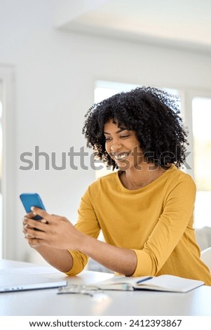 Happy smiling young African American woman customer sitting at home table looking at cellphone while holding mobile cell phone in hands texting using smartphone shopping online in application.