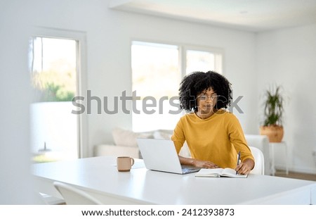 Young pretty African American woman entrepreneur or student using laptop computer sitting at home table in living room interior learning or working with ebusiness online, elearning and writing notes.
