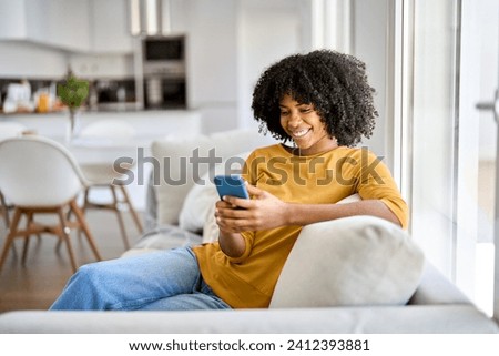 Happy smiling young African American woman customer sitting at home on sofa looking at cellphone holding mobile cell phone in hands texting using smartphone scrolling media, buying online. Authentic.
