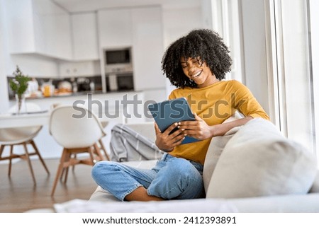 Smiling young African American woman using digital tablet relaxing on couch at home. Happy lady sitting on sofa looking at tab laughing holding pad in hands watching funny video in living room.