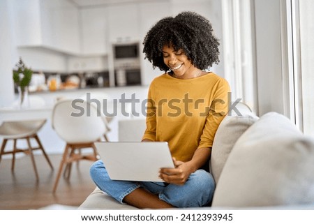 Happy young African American woman sitting on couch at home using laptop. Smiling ethnic lady relaxing on sofa looking at computer technology device browsing web buying online doing shopping.