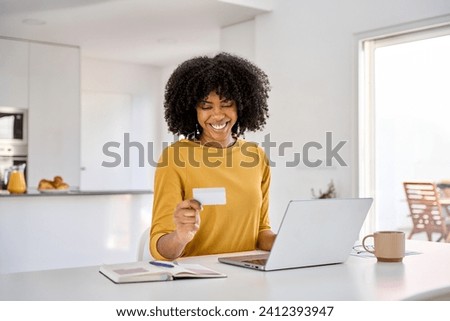 Young adult smiling happy African American woman holding credit card using laptop computer paying or buying online making ecommerce shopping purchase sitting at home table in kitchen. Royalty-Free Stock Photo #2412393947