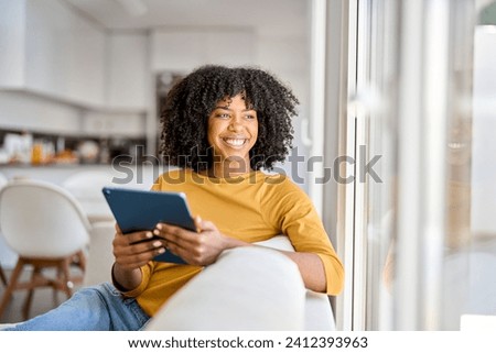Happy African American lady using tab device looking away on sofa at home. Smiling pretty young woman sitting on couch relaxing looking away at window holding digital table in kitchen. Royalty-Free Stock Photo #2412393963
