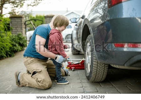 Cute toddler boy helping his father to change car wheels at their backyard. Father teaching his little son to use tools. Active parent of a small child. Royalty-Free Stock Photo #2412406749