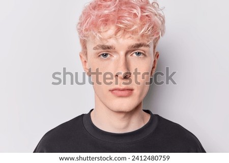 People emotions concept. Indoor photo of young calm relaxed European guy with pink hair wearing black t shirt standing in centre on white background looking straight at camera keeping hands down