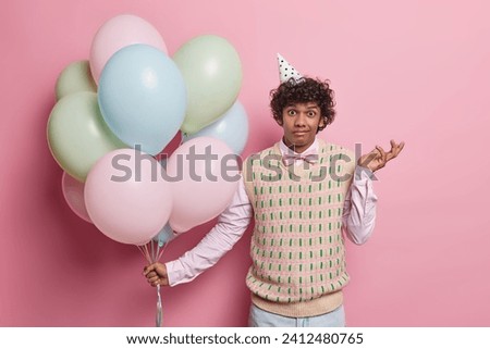 Hesitant birthday man shrugs shoulders with clueless expression holds bunch of inflated colorful balloons doesnt know why guests didnt come isolated over pink background. Celebration concept