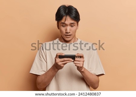 Waist up shot of brunet Japanese man holds smartphone horizontally plays video games tries to pass difficult level stares impressed at screen dressed in casual t shirt isolated over brown background