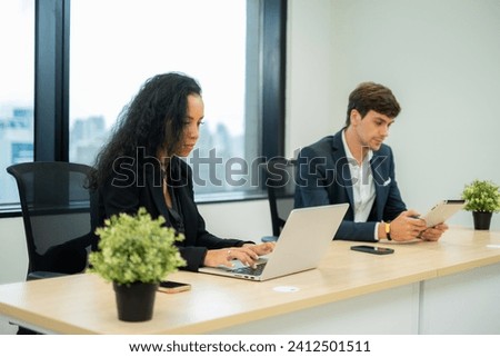 White working woman, employee, business owner sitting at desk using laptop while her co-worker using tablet at office on building with skyscaper view.