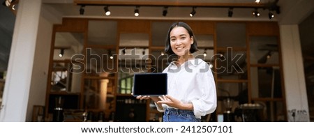 Smiling asian woman showing digital tablet screen, cafe owner showing smth, standing in front of cafe entrance.