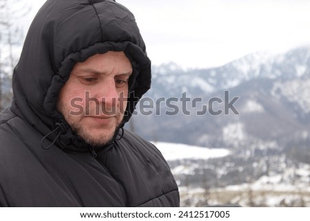A man in a black jacket and a hood pondered against the background of snow-capped mountains