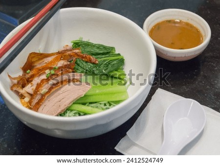 Close-up of dried green noodles with roasted duck in a white bowl with sliced green onions. Boiled bok choy with a white spoon, two colored chopsticks, and a small bowl of sauce, Chinese food Royalty-Free Stock Photo #2412524749