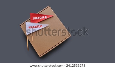 Small paper flags with the warning 'Fragile' as a label and empty cardboard box on monochrome gray background. The concept of shipping fragile products. Copy space
