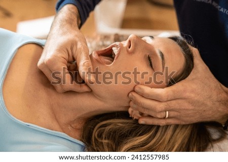 An osteopath performs a jaw adjustment on a female patient, focusing on alleviating tension and improving joint function. Royalty-Free Stock Photo #2412557985