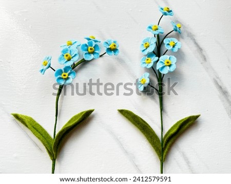 Two stems of light blue forget-me-not flowers made of felt fabrics on a white marble motif background, totally handmade. All parts are cut by free hand using scissors, without any pattern
