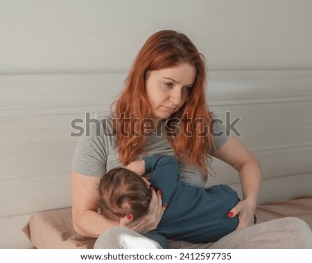 An unhappy woman breastfeeds her son and sits on the bed. Postpartum depression. Royalty-Free Stock Photo #2412597735