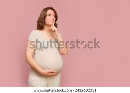 A pregnant woman with her finger on her lips on a pink studio background. Pregnancy in a woman with a belly showing a sign of silence, copy space