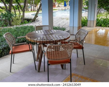 A coffee table set with one circular wooden table and four simple armchairs set up at a clean terrace overseeing a tidy garden by the beach and pool.
