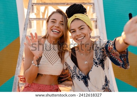 two stylish smiling beautiful women relaxing having fun on beach in summer fashion trend outfit, colorful apparel boho style, having tropical vacation using smart phone taking selfie photo on camera