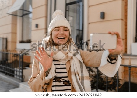 stylish young pretty woman walking in winter street wearing beige coat, knitted hat, scarf, smiling happy cold season fashion trend, taking selfie, making photo on phone
