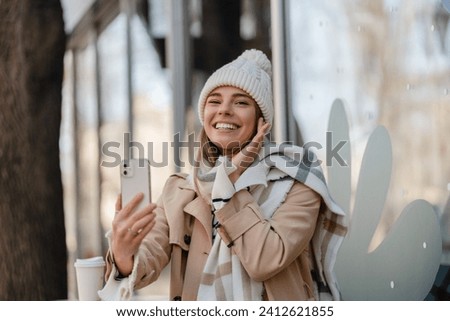 stylish woman walking in winter street wearing beige coat, knitted hat, scarf, smiling happy cold season fashion trend, taking selfie, making photo on phone