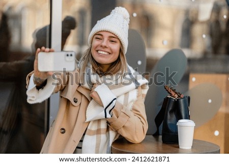 stylish woman walking in winter street wearing beige coat, knitted hat, scarf, smiling happy cold season fashion trend, taking selfie, making photo on phone