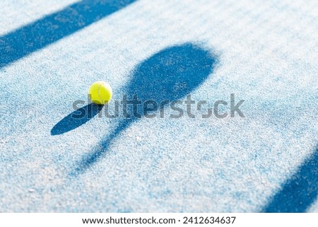 Photo of a padel ball and the shadow of a racket on the floor of an outdoor court. Padel concept. Blue outdoor padel court.