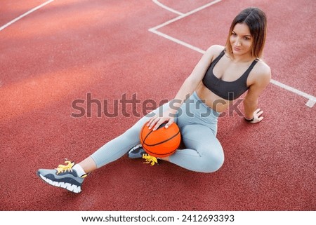 High angle view of woman in sportswear taking a workout break, sitting on an outdoor basketball court, holding a ball and relaxing