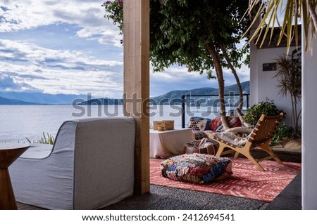 The image depicts a relaxing lakeside outdoor seating area with comfortable furniture, colorful cushions, and a stunning view of the lake and mountains, creating an atmosphere of peaceful retreat.