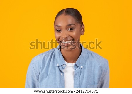 Headshot of pretty African American woman in denim shirt, confidently smiling over yellow studio backdrop. Modern fashion and female beauty concept, portrait for advertisement banner
