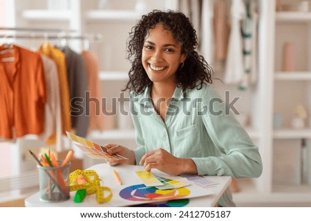 Happy creative fashion designer working with color swatches and pie charts in her bright studio, surrounded by trendy wardrobe, smiling at camera