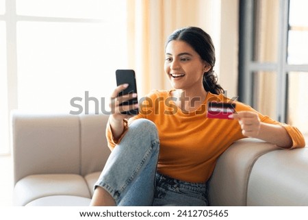 Smiling young indian woman enjoying online shopping on her smartphone at home, happy eastern female holding credit card, sitting comfortably on couch in living room, making purchases, copy space
