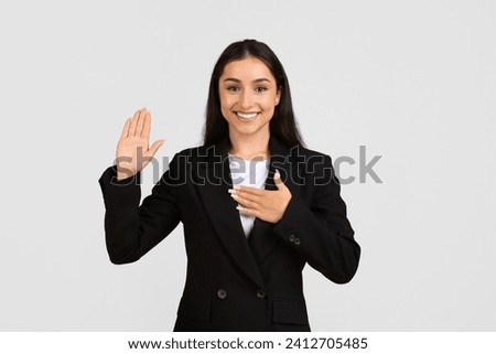 Sincere european businesswoman making pledge or swearing an oath with her right hand raised and left hand on her chest on grey background Royalty-Free Stock Photo #2412705485