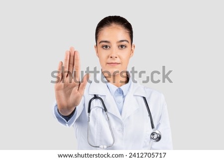 Serious young european doctor with a stethoscope gesturing 'stop' with her hand, emphasizing the importance of health safety measures and boundaries, isolated on gray studio background