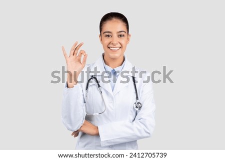 A positive european lady doctor wearing a lab coat and a stethoscope around her neck makes an okay sign, signifying satisfaction and good health outcomes, isolated on gray studio background