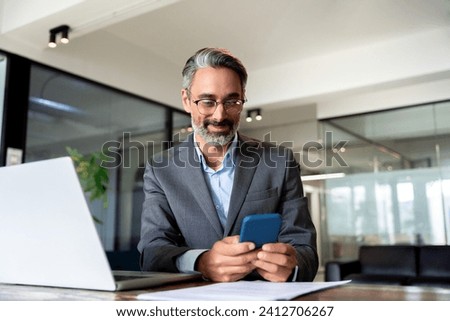 Middle aged Hispanic business manager ceo using cell phone mobile app, laptop. Smiling Latin or Indian mature man businessman holding smartphone sit in office working online on gadget with copy space. Royalty-Free Stock Photo #2412706267