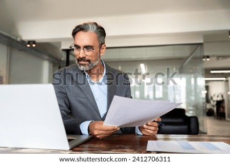 Focused Hispanic businessman with paperwork in office. Busy Latin or Indian male business man accountant analyst holding documents, work at laptop computer doing online trade market tech research.  Royalty-Free Stock Photo #2412706311