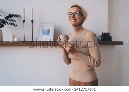 Positive middle aged woman smiling with coffee cup and relaxing at home in cozy room near shelf with photo frame while looking away