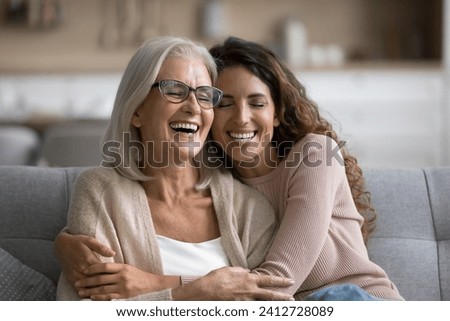 Happy excited senior mom and adult daughter woman having fun at home, hugging with heads touch, sitting on sofa, laughing with closed eyes, enjoying funny talk, leisure, family affection Royalty-Free Stock Photo #2412728089