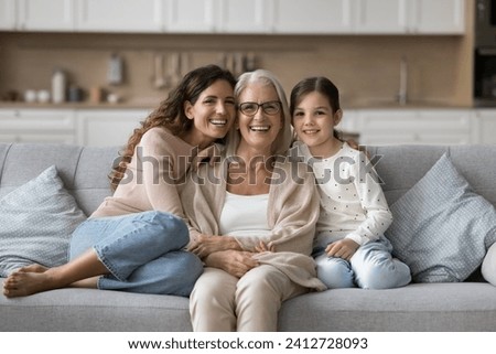 Happy grandma, mom and granddaughter kid sitting close on home sofa. Young mother and girl hugging grandmother with love, affection, laughing, smiling. Three female generations portrait Royalty-Free Stock Photo #2412728093