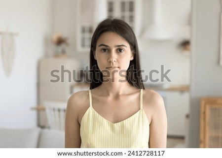 Head shot of attractive young 25s single confident woman pose in cozy domestic room. Portrait of beautiful millennial female staring at camera looks serious, spend time at own or rented dwelling alone
