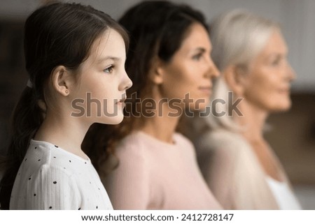 Side shot of serious little tween child girl standing in row with mom and grandmother in blurred background, looking forward away, posing for family portrait of three female generations