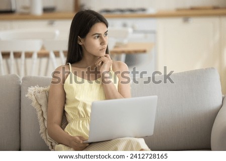 Serious, deep in thoughts young female sits on sofa with laptop, consider answer to client by e-mail, think on solution, studying or working on-line, learn new software, faced up with difficult task Royalty-Free Stock Photo #2412728165