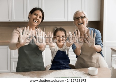 Cheerful grandmother, happy mother and excited little girl in aprons showing floury hands at camera, posing for portrait, laughing, baking in home kitchen, having fun, enjoying family cooking Royalty-Free Stock Photo #2412728193