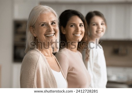 Happy attractive blonde senior grandma standing in row with adult daughter woman and little granddaughter kid, looking at camera with toothy smile, posing for three generations family portrait Royalty-Free Stock Photo #2412728281